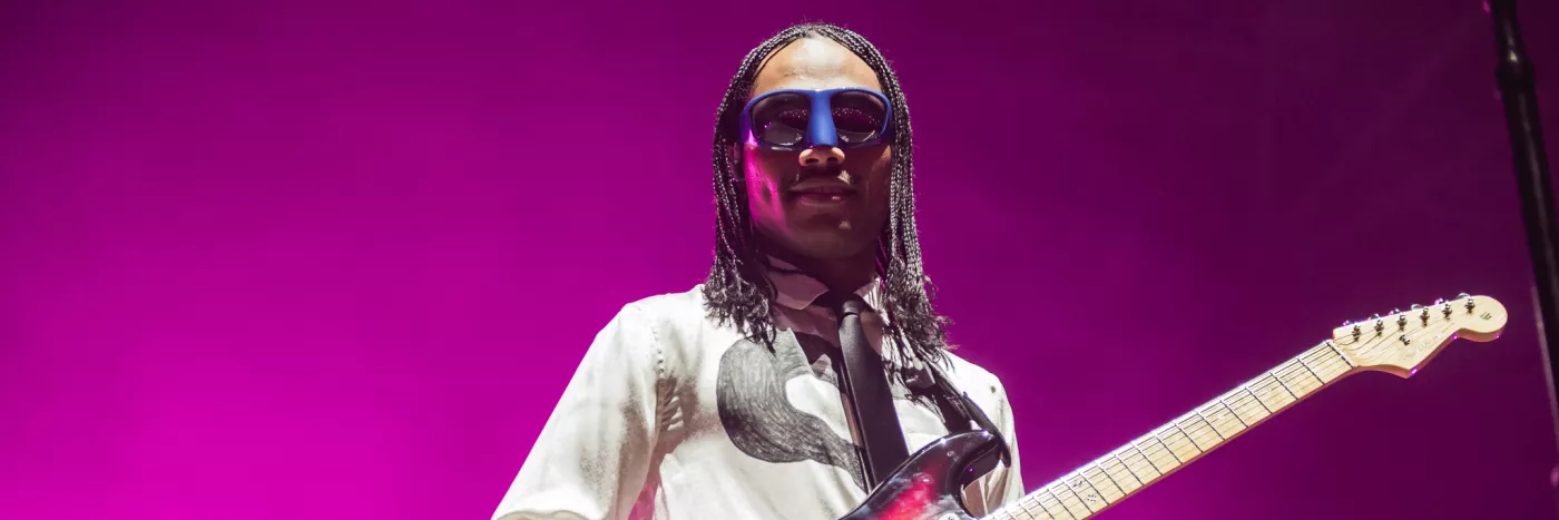 A person wearing googles and a white long sleeve t-shirt playing a guitar against a bright purple background.