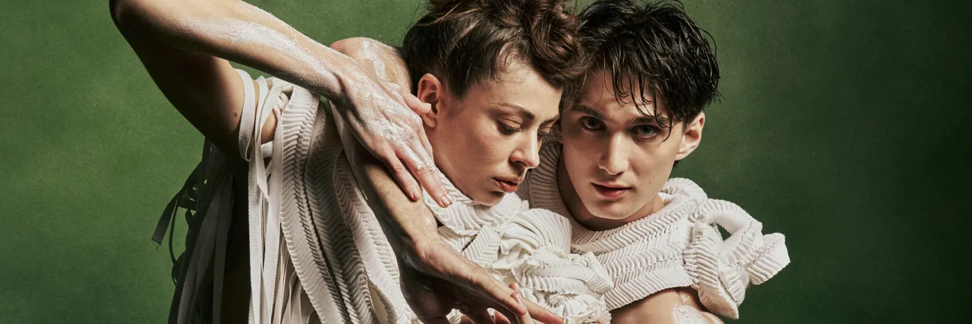 Two dancers in off-white textured costume with arms interlocked and white clay on their bare arms, against a dark green background.