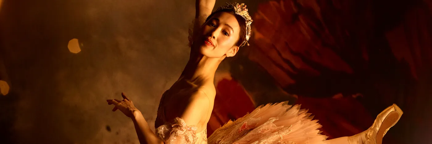 A ballerina in a pink jewelled tutu and tiara mid-pose, against a dark red and grey background.