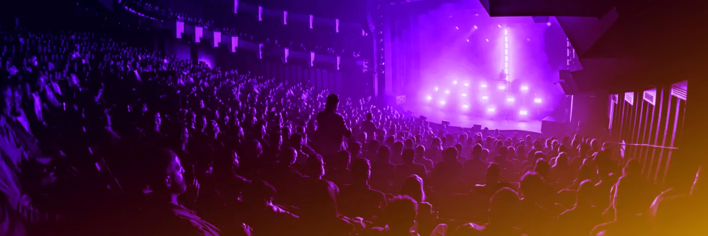 A crowd of people in a concert hall with an artist on stage surrounded by lights.