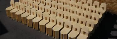 A group of 60 cream chairs with a square cut out in the back sit angled to the left in the sun infront of a brown bricked wall.