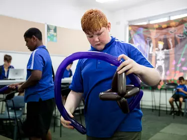 A boy with red hair holding a shaped balloon in a classroom.